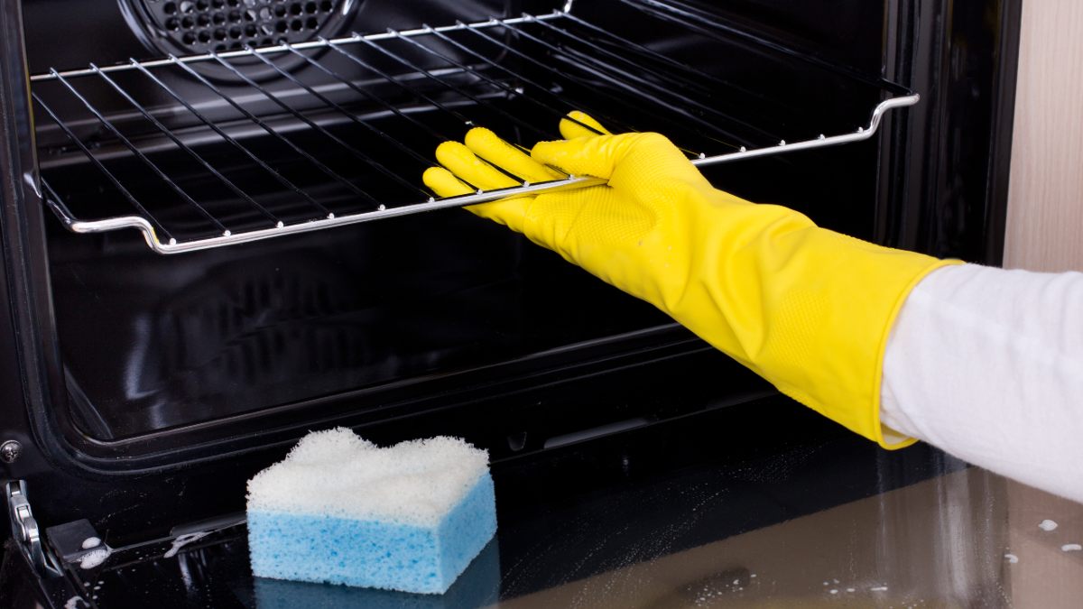 A hand wearing a yellow rubber glove reaches into an oven to grab a wire rack. A sponge sits on the oven's open glass door
