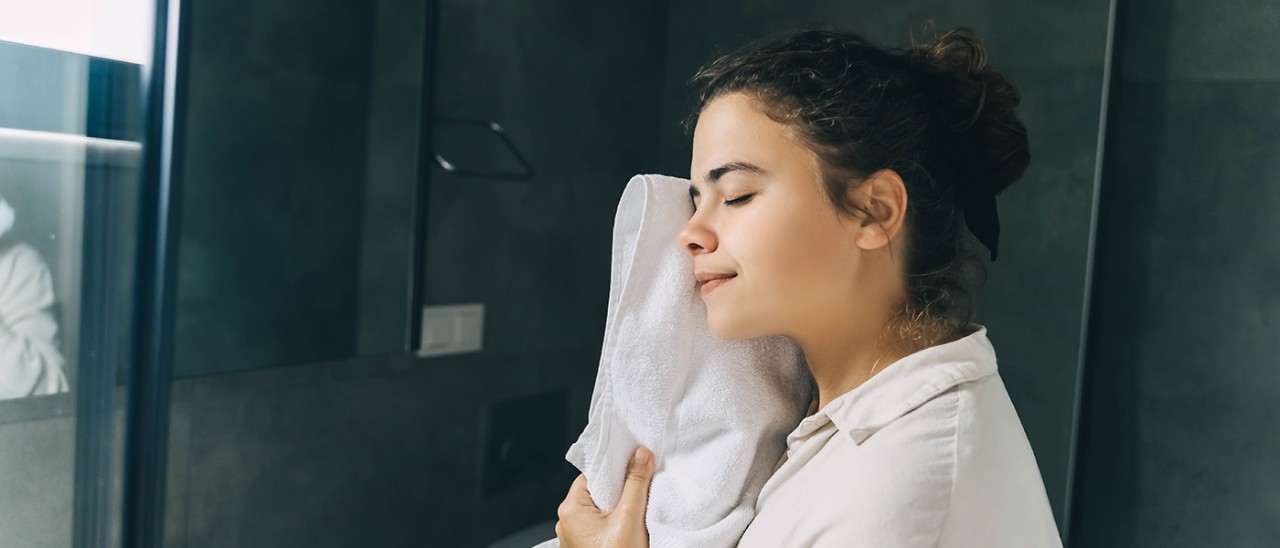 Woman in bathroom holding warm towel against her face.