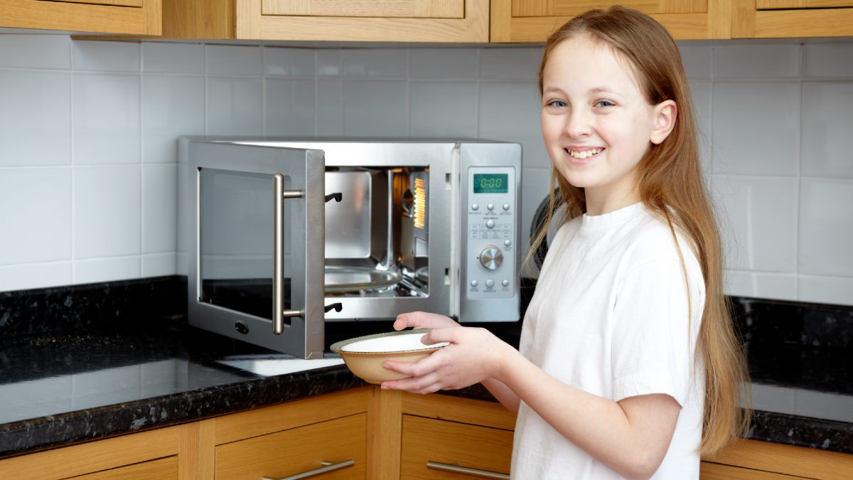 young girl holding bowl in front of open microwave in kitchen