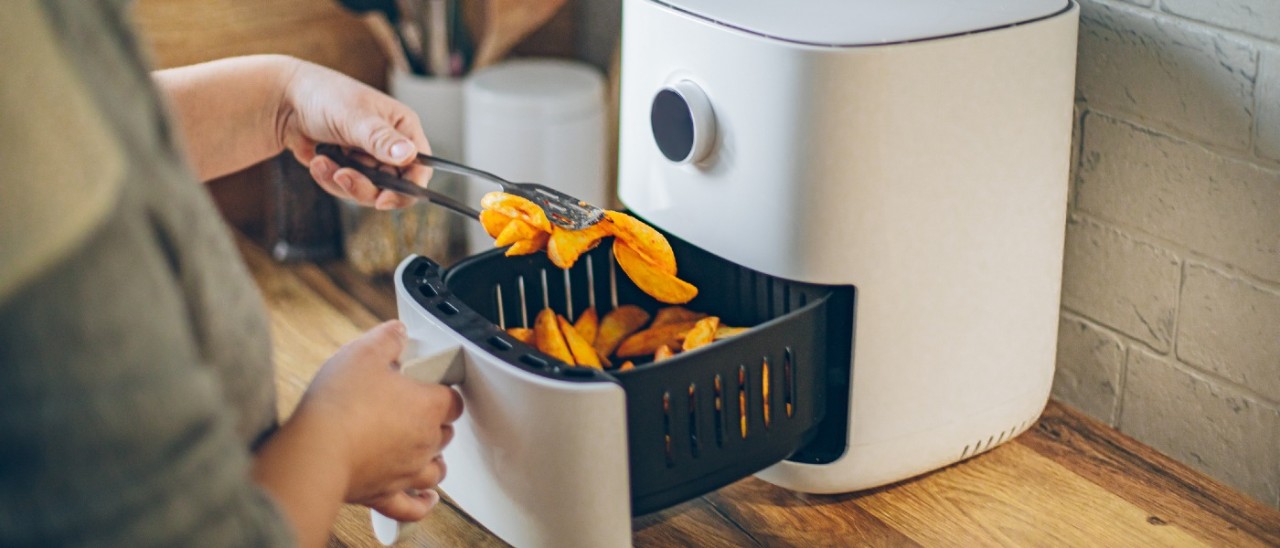 A person lifting potato wedges out of a white air fryer on a rustic kitchen bench