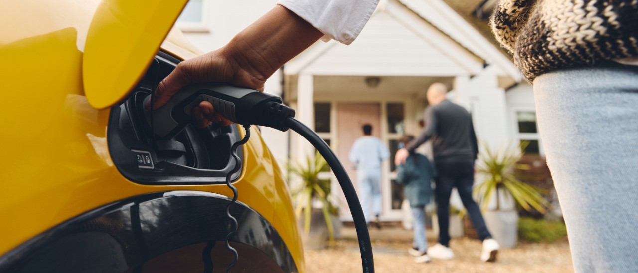 A close-up of a person putting a charging cable into their bright yellow EV. In the background a man and two children can ne seen walking towards a front door of a house