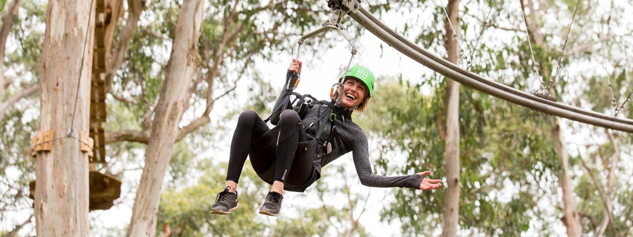 A woman strapped into a harness and wearing a helmet as she gleefully swings on a zip line through a eucalypt forest