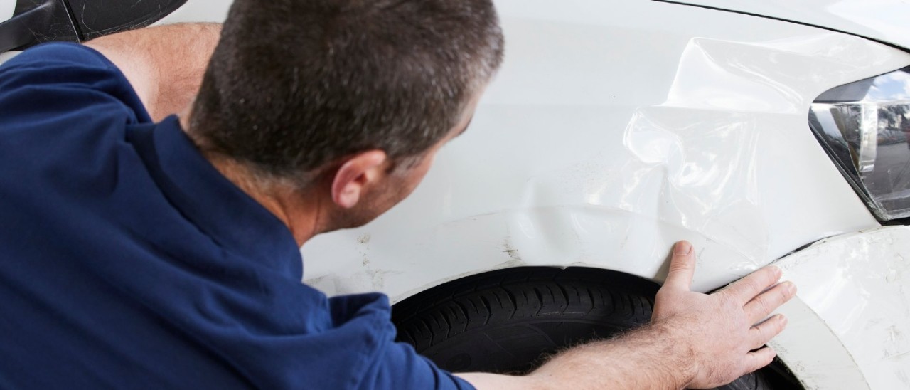 A panel beater inspects a dent on the front bumper of a white sedan-type car