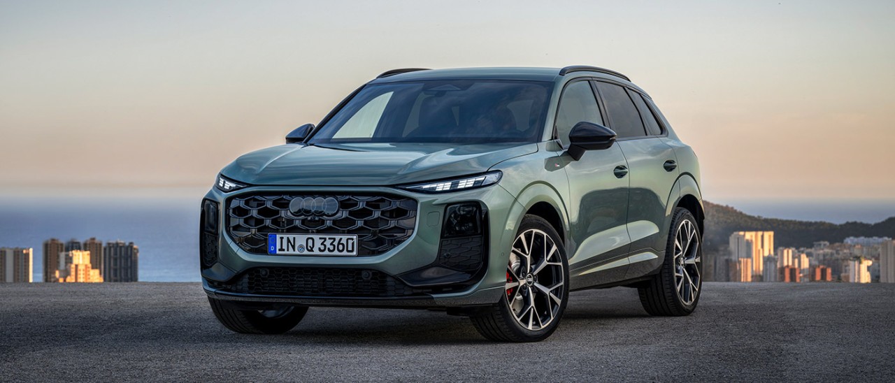 Sage green metallic Audi Q3 small SUV at dusk on coastline in Europe overlooking mountain, city and sea.
