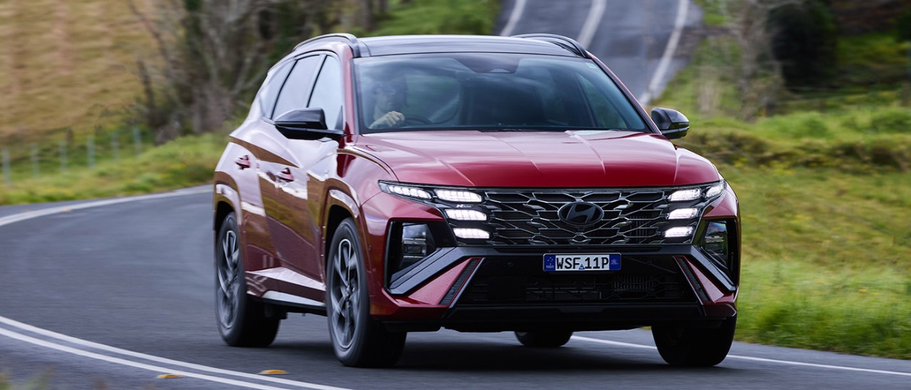 2025-Hyundai-Tucson-Premium-N-Line-Hybrid-banner-1400x600 Red Hyundai Tucson Premium N Line Hybrid SUV taking a corner on country road.