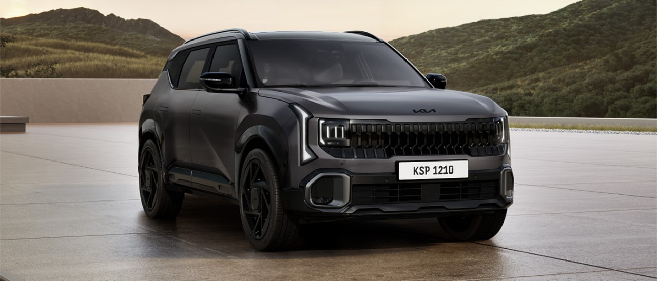 Dark grey Kia Seltos X-Line SUV parked on large concrete forecourt in front of green highland mountains.