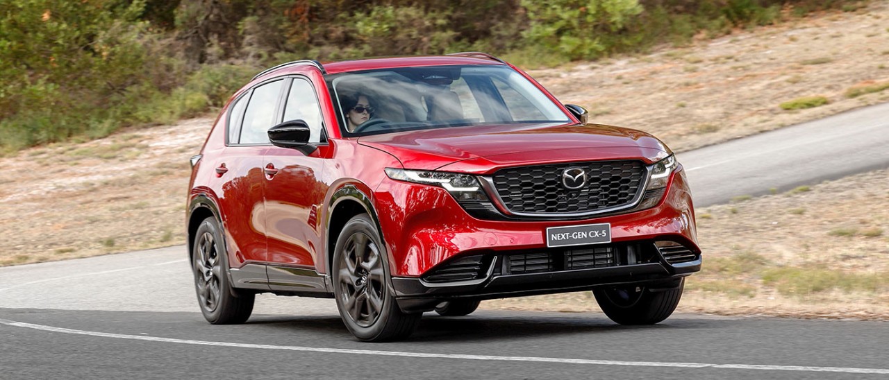 Woman with dark hair and sunglasses driving red Mazda CX-5 SUV through corner at bottom of steep hill at Lang Lang proving ground in Victoria.