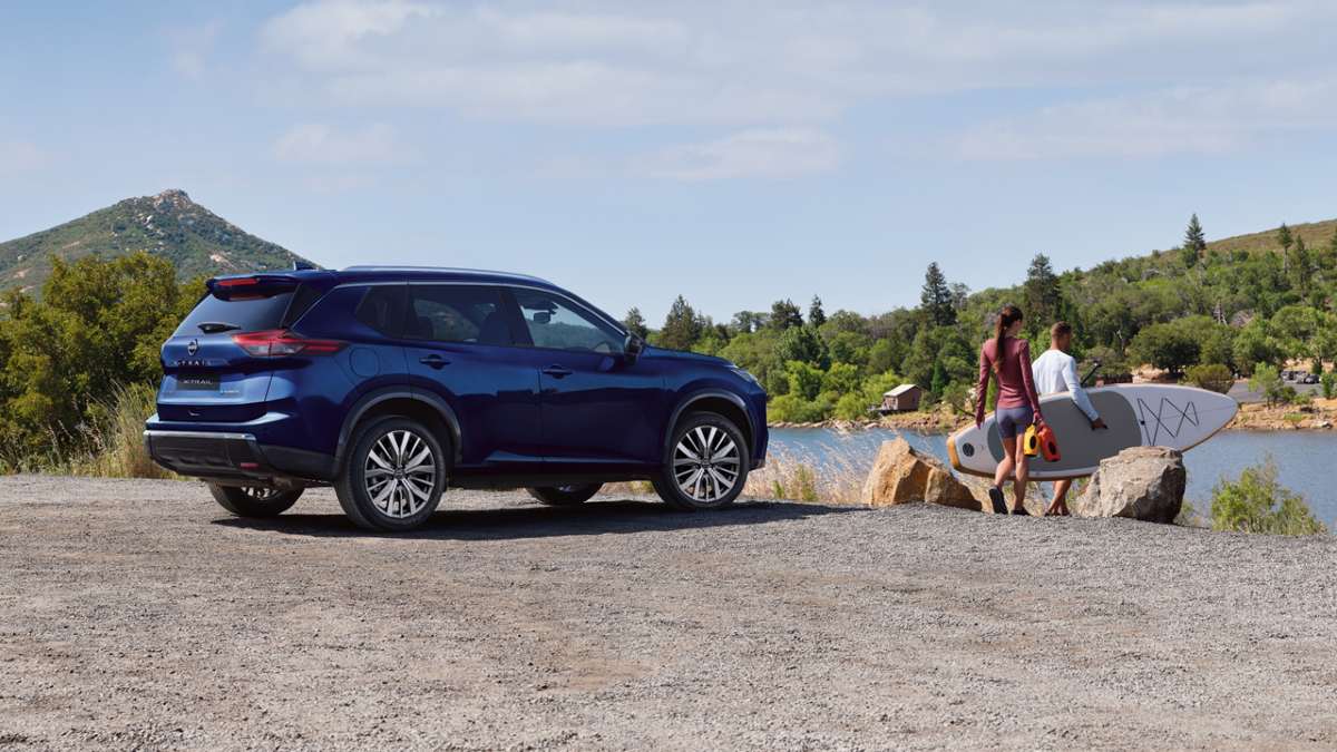 dark blue 2026 Nissan X-Trail parked on gravel by a river; people are carrying a paddleboard in