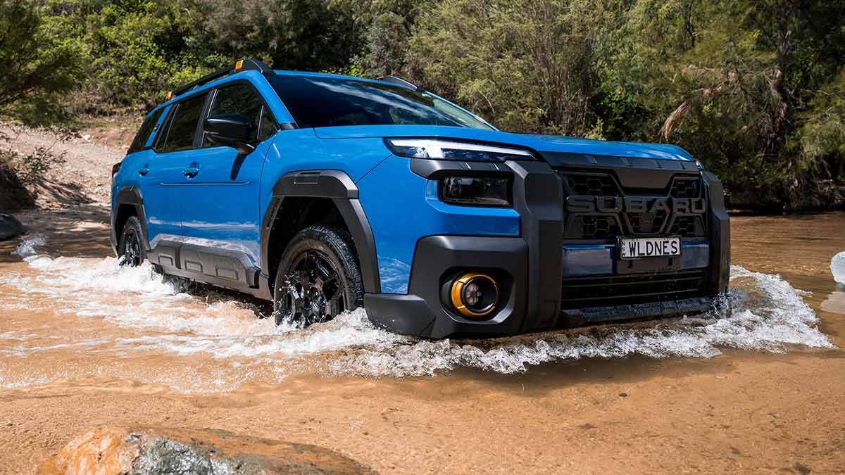 Blue Subaru Outback Wilderness SUV crossing shallow creek with light gravel sand and rocks at side in Australian bush.