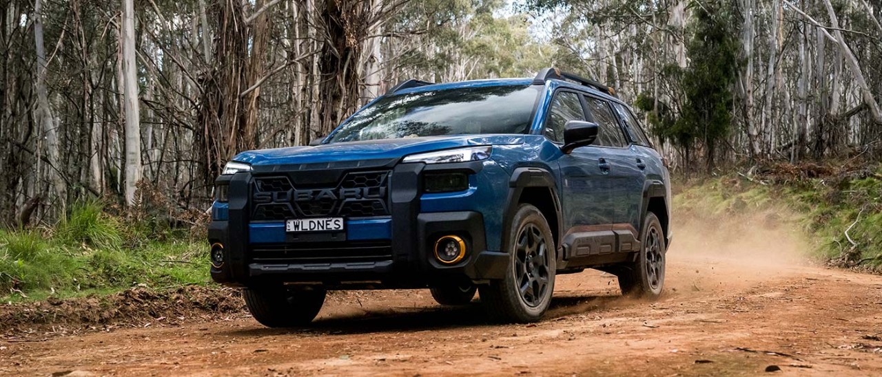 Blue Subaru Outback Wilderness SUV driving on dusty red dirt road lined with gum trees in regional Australia.