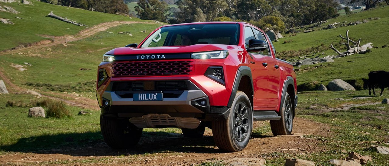 Red Toyota HiLux Rogue ute on hilly, rocky off-road trail with mountain range in distance.  