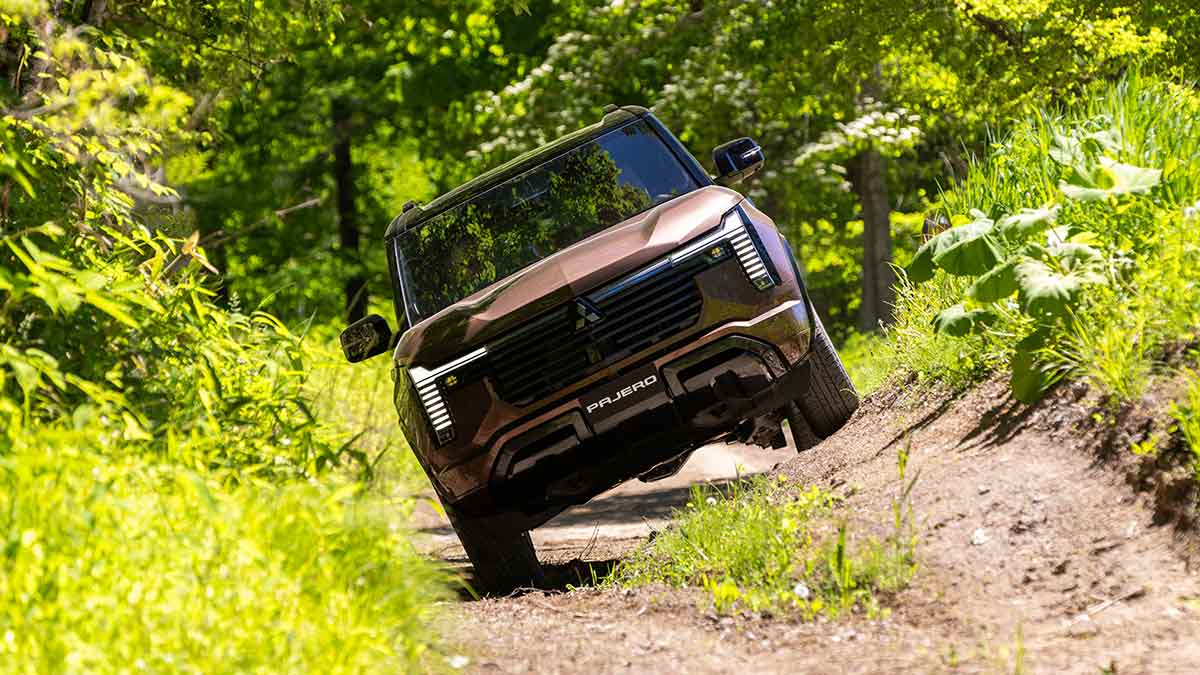 Front-on shot of bronze Mitsubishi Pajero SUV tilted on off-road track in jungle setting.