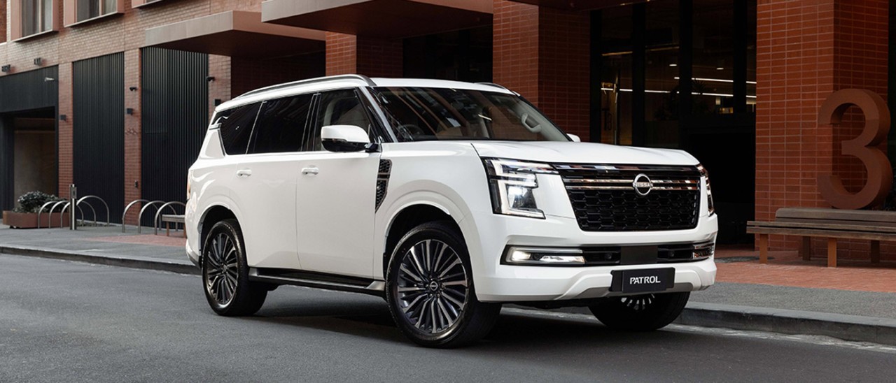 A white Y63 2027 Nissan Patrol SUV parked on a street in front of a red brick industrial office complex.