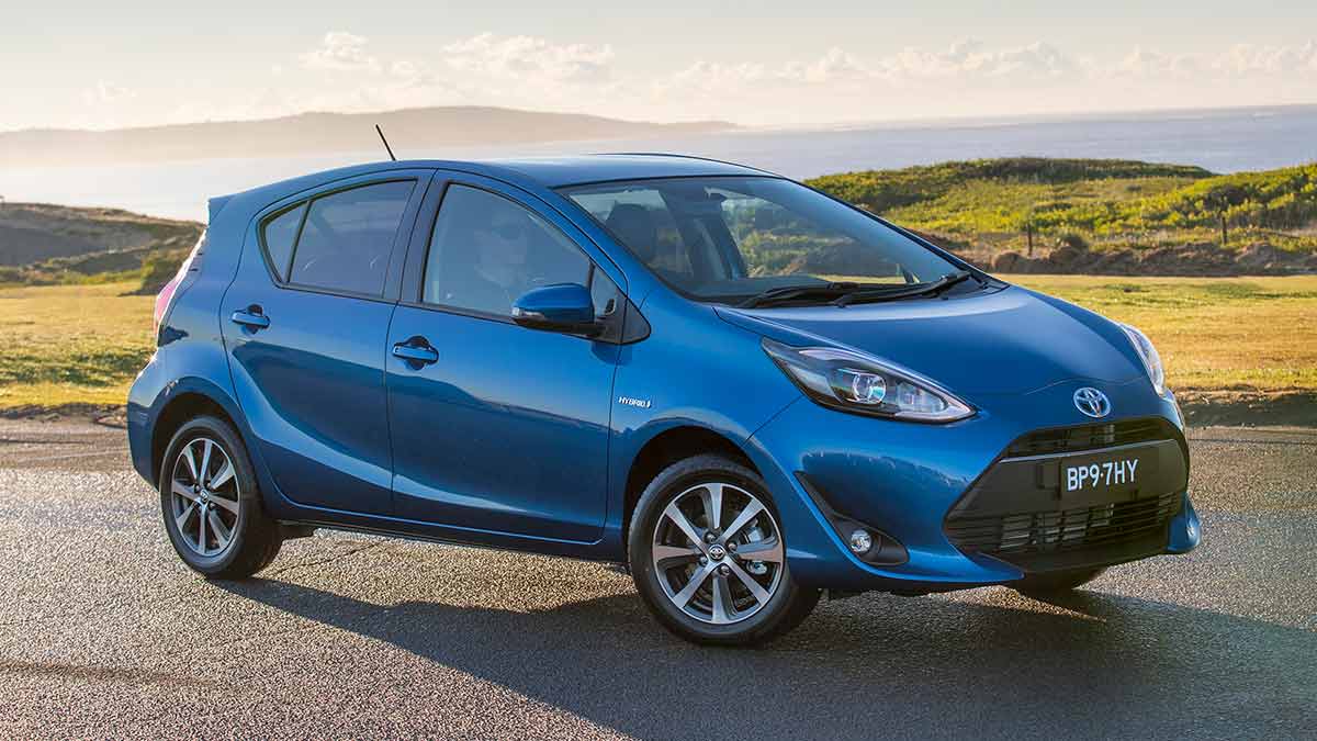Blue 2017 Toyota Prius C small hybrid hatch at beachside carpark with ocean and headland behind.