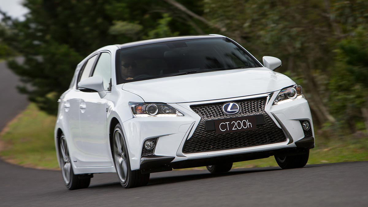 White Lexus CT200h small car on flowing bitumen country road bordered by pine trees and bush.