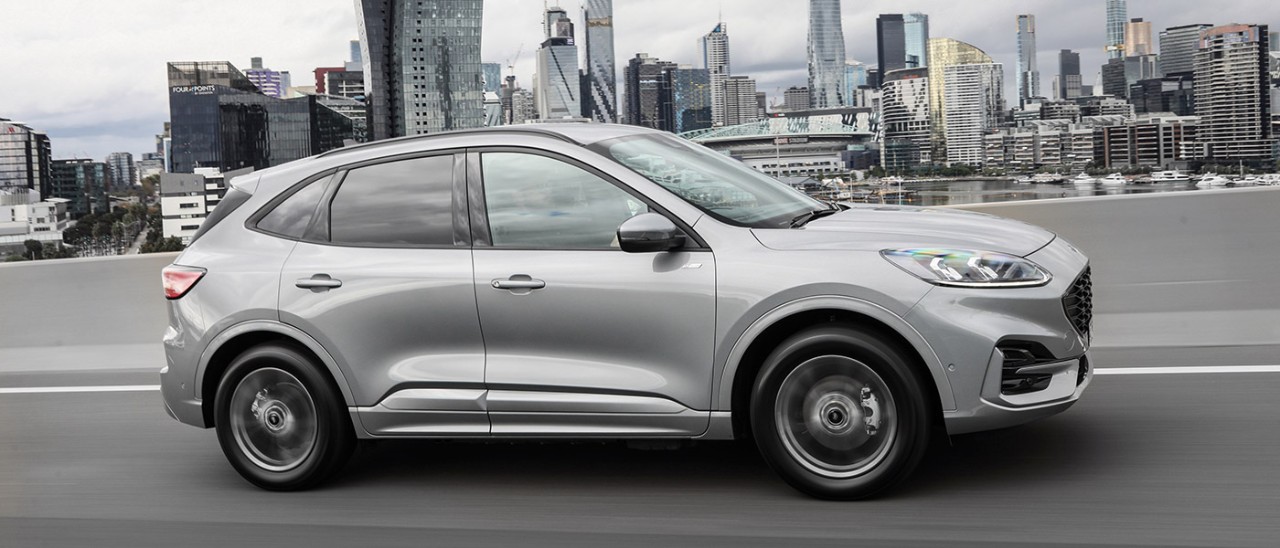 Silver Ford Escape PHEV SUV driving on Bolte Bridge in Melbourne with Docklands and city CBD behind.