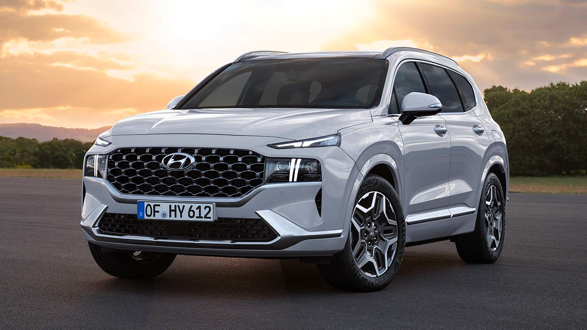 White Hyundai Santa Fe Hybrid SUV parked on large forecourt in front of bush and lowering sun shining brightly.