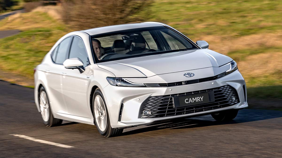 Man driving white Toyota Camry Ascent Sport sedan on winding country road with patchy green-brown grass on verge.