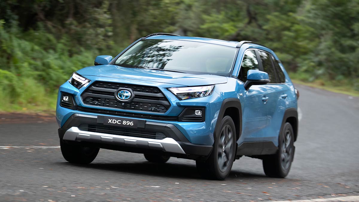 Light blue Toyota RAV4 Hybrid SUV driving through corner in lush green bush setting.