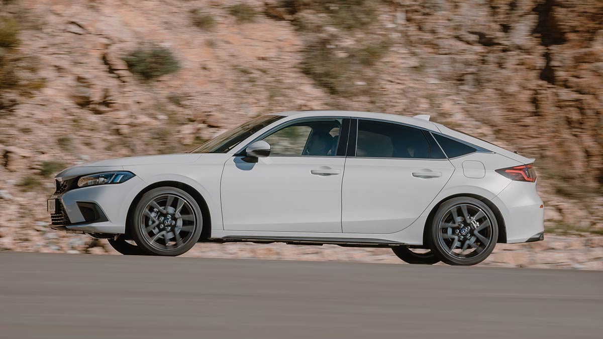 Side-on profile shot of white Honda Civic hybrid sedan driving on brown-paved road next to high rocky bank.