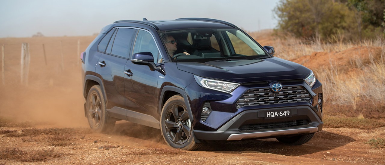 Man with sunglasses driving black Toyota RAV4 Hybrid SUV on dusty red dirt road next to dry agricultural land.