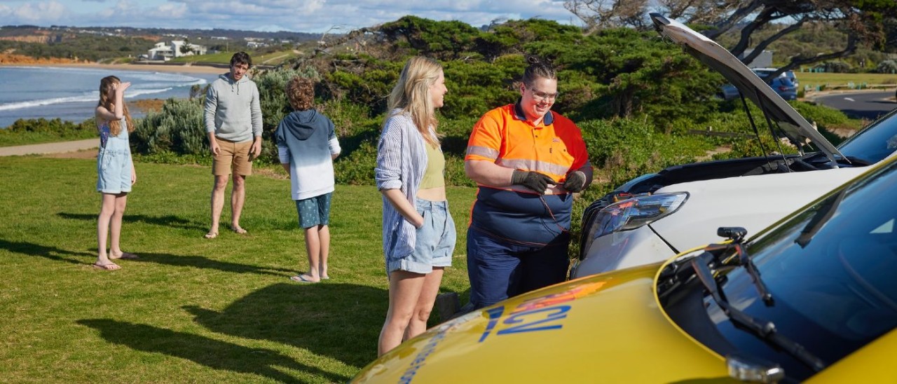 A woman watches on happily as an RACV Emergency Roadside Assistance technician works on their vehicle