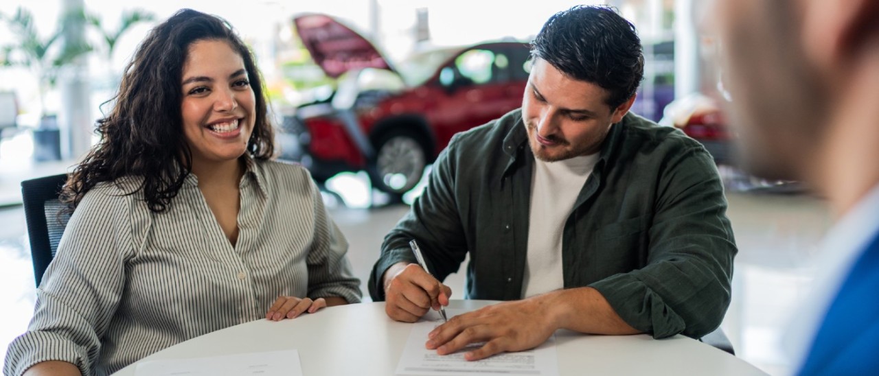 man signing finance documents while female partner smiles; lender in foreground; red car in background