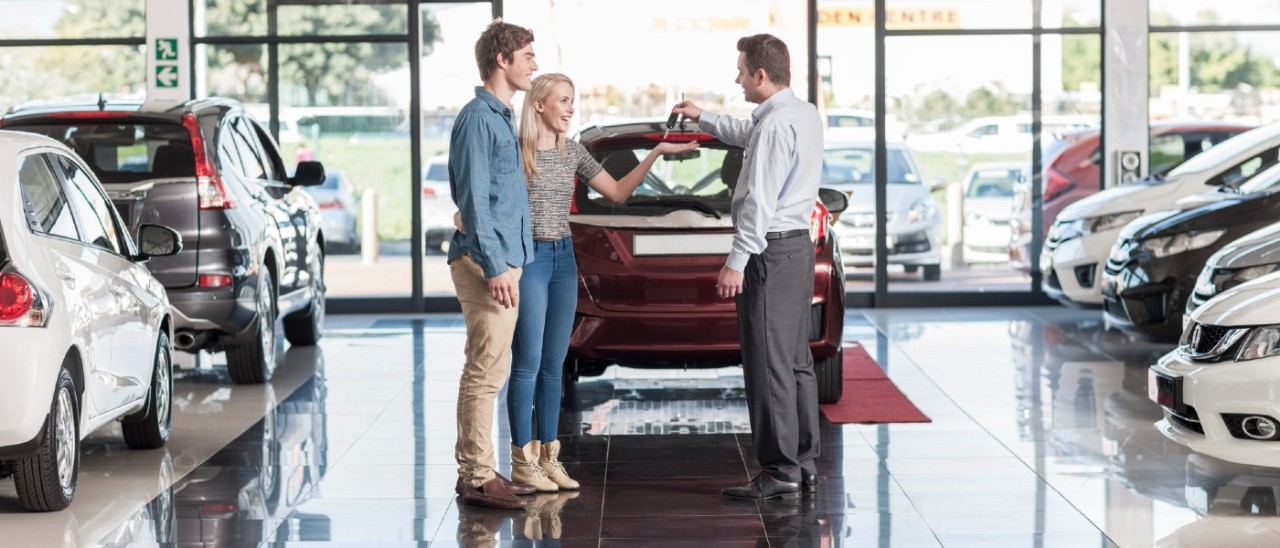 car salesman handing keys to young couple in front of small red car