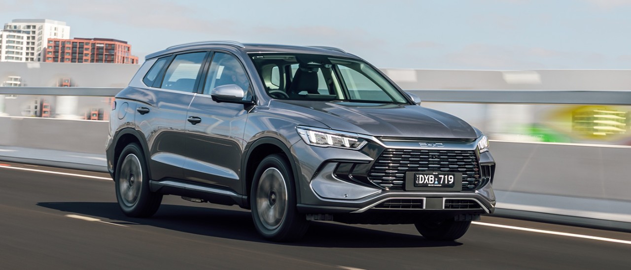 Metallic grey BYD Sealion 5 medium SUV with Victorian number plates driving on city overpass with tower blocks in background.