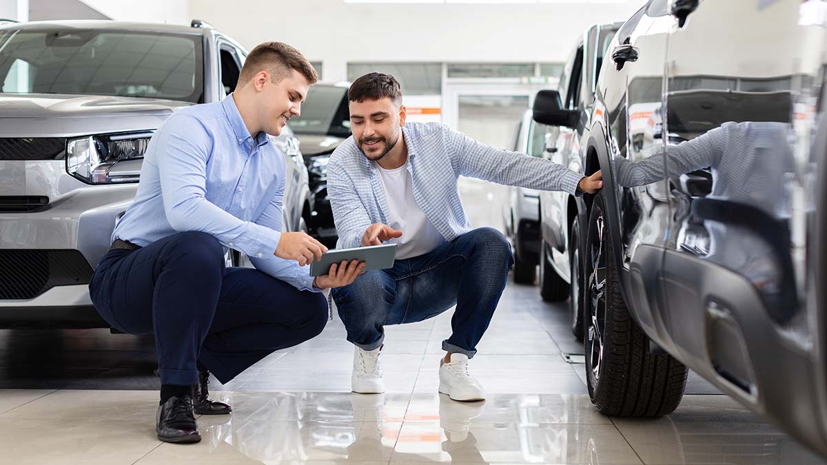 A salesman and car buyer kneel beside a car while looking at a hand-held computer in a dealership.