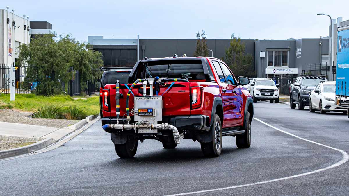 Rear view of red Toyota HiLux ute rigged up with fuel economy and emissions testing equipment, driving through light industrial area.