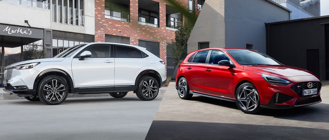 Small-SUV-vs-Hatch-Honda-HR-V-Hyundai-i30-banner-1400x600 Split image of white Honda HR-V Hybrid small SUV on city street and red Hyundai i30 N Line hatchback in factory carpark.