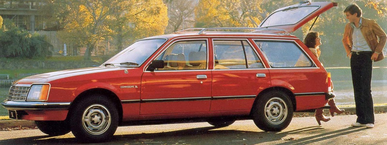 Two people conversing at the back of a red Commodore VB station wagon