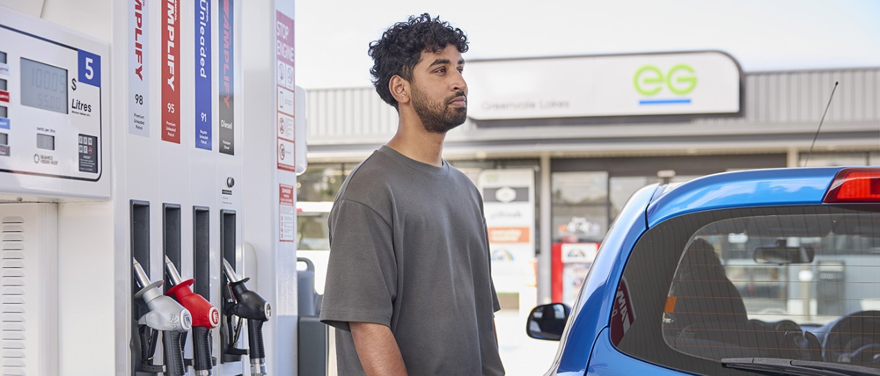 Bearded man with grey t-shirt looking into distance while filling up blue car at petrol station bowser.