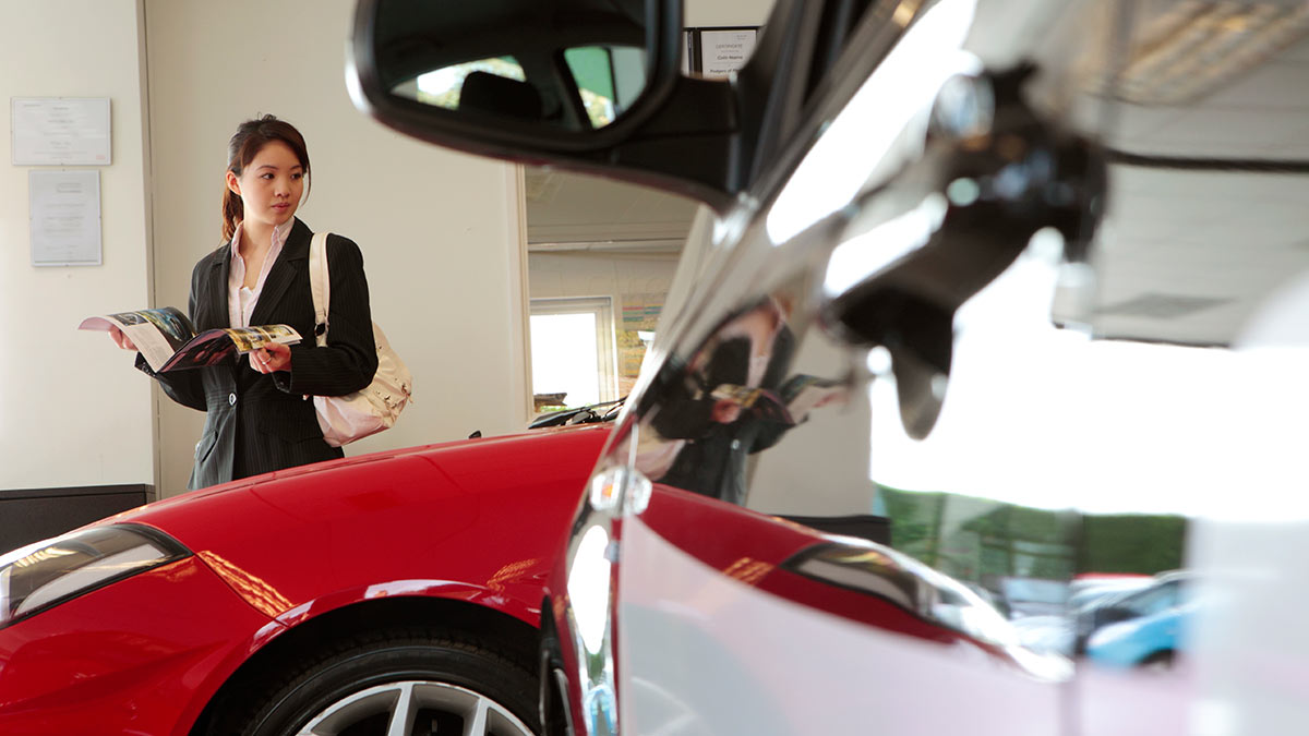 a woman holding a brochure walks around a car dealership.