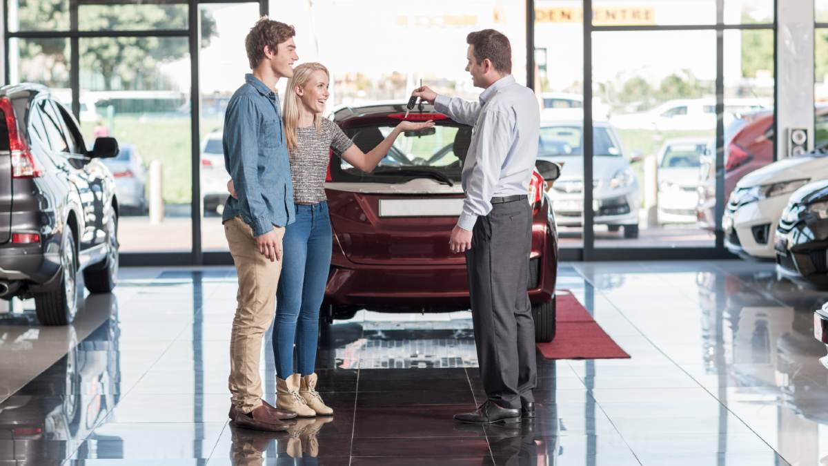 car salesman handing keys to young couple in front of small red car