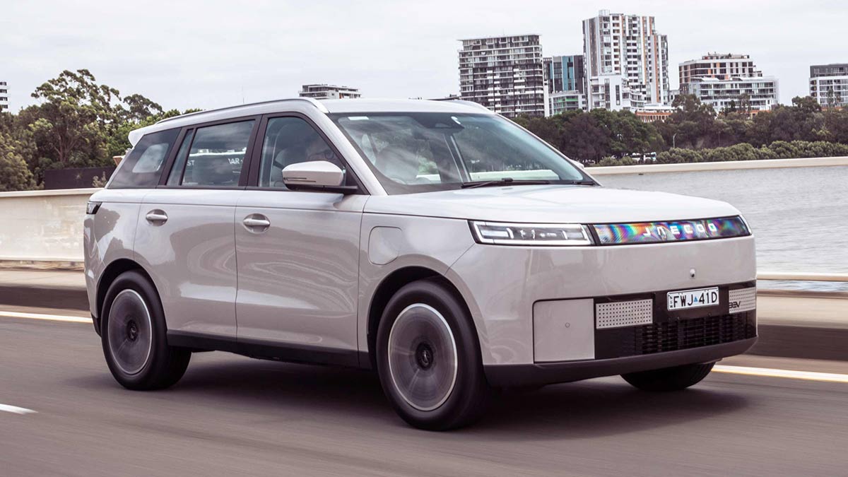 White Jaecoo J5 EV on dual-carriageway road crossing waterway with parkland and city towers behind.