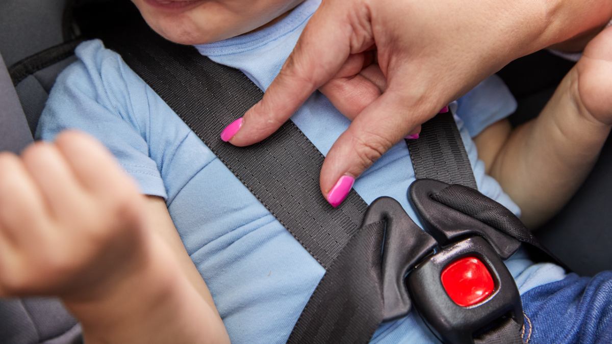 A person doing the pinch test on a child car seat straps while a toddler sits in the seat