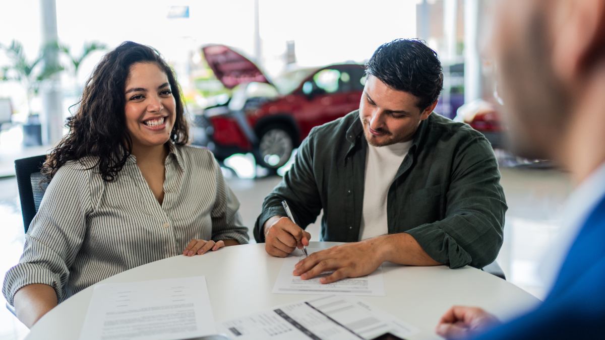 man signing finance documents while female partner smiles; lender in foreground; red car in background