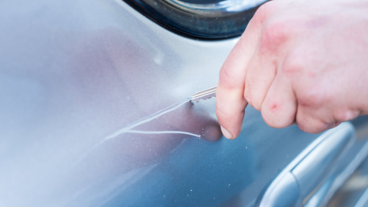 Close-up of hand with key making deep scratches into blue car's body panel at back door