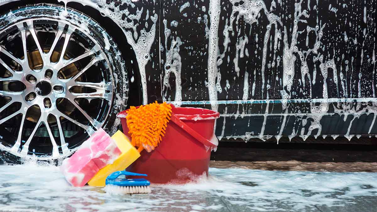 Black car covered in soap suds with red bucket in foreground with pink and yellow sponges, orange towel and blue brush