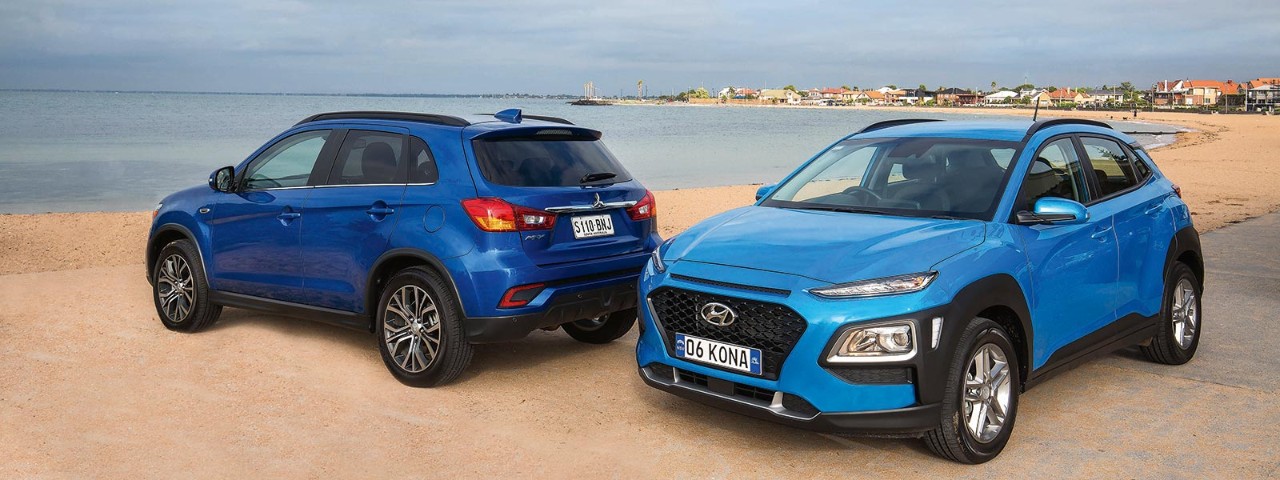 mitsubishi-vs-hyundai-1600x600.jpg A dark blue Mitsubishi ASX XLS parked next to a light blue Hyundai Kona Active at alternating angles at the beach