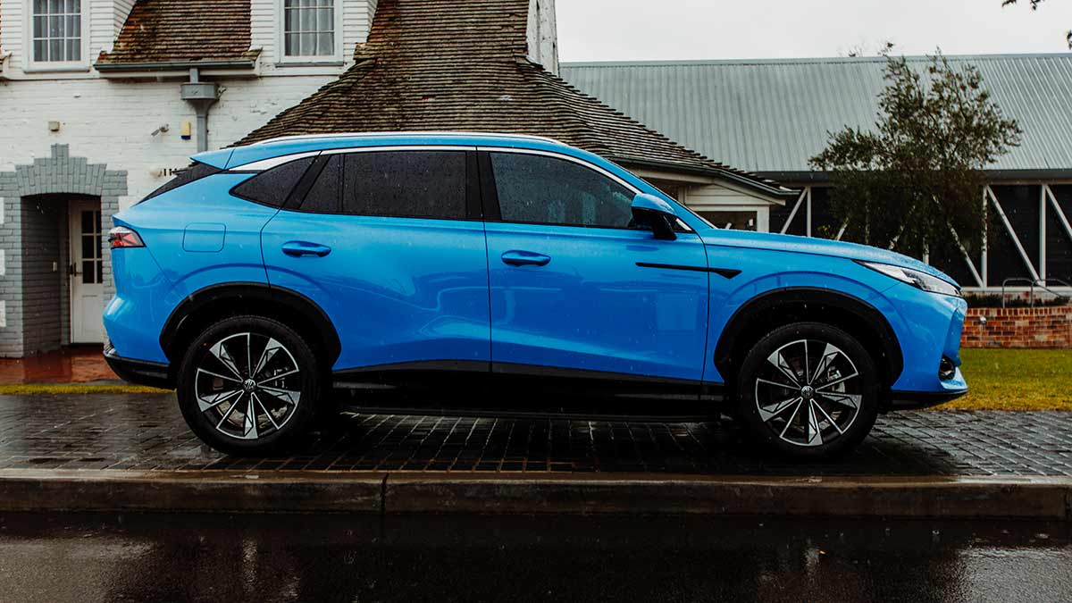 Side view of blue MG HS medium-size hybrid SUV on wet pavement in front of white-brick heritage house.