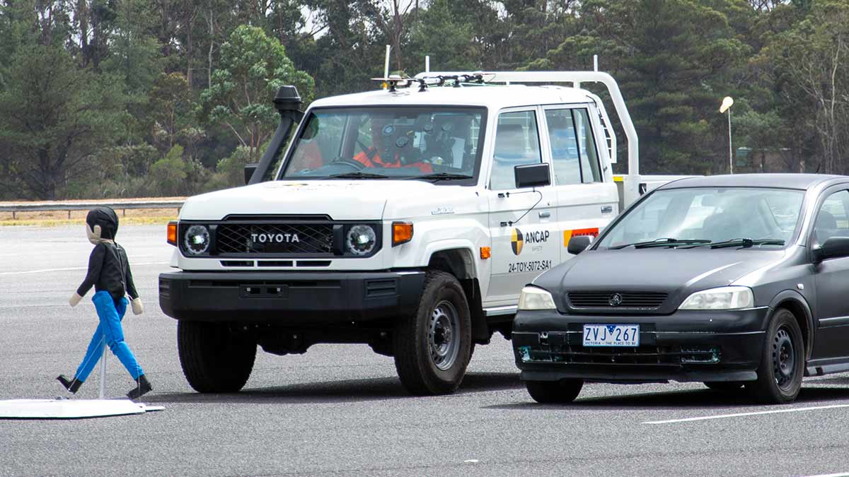 White Toyota LandCruiser 70 Series ute next to black Holden car with child pedestrian dummy walking in front during ANCAP safety test.