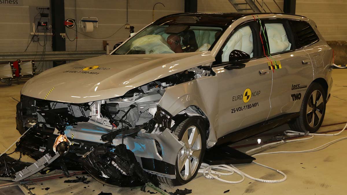 Silver Volvo EX90 SUV under yellow light in crash lab with severely damaged front end after frontal offset Euro NCP test.