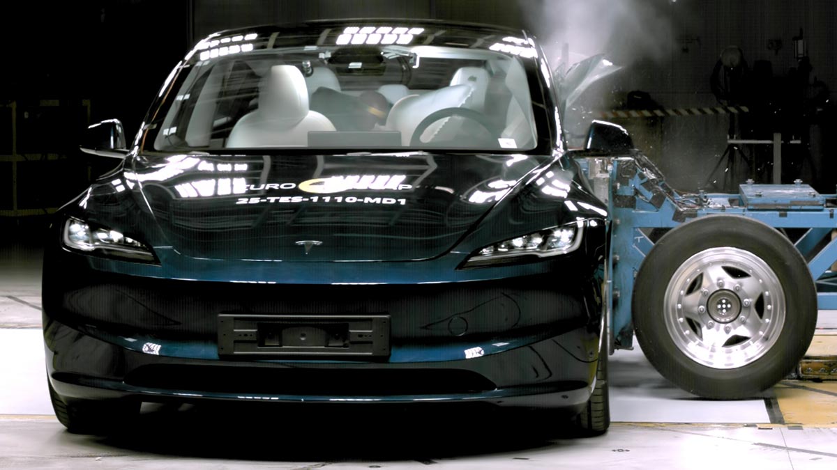Dark metallic blue Tesla Model 3 EV sedan being hit side-on by a deformable moveable barrier in Euro NCAP testing.