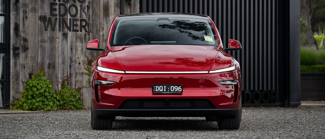 Red Tesla Model Y SUV parked in front of Mt Macedon winery building with grey weathered boards and black steel door.