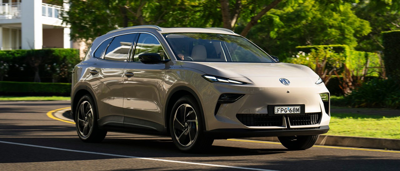 Champagne gold metallic MG S5 EV small SUV in leafy green suburban street with white house and hedgerows behind.