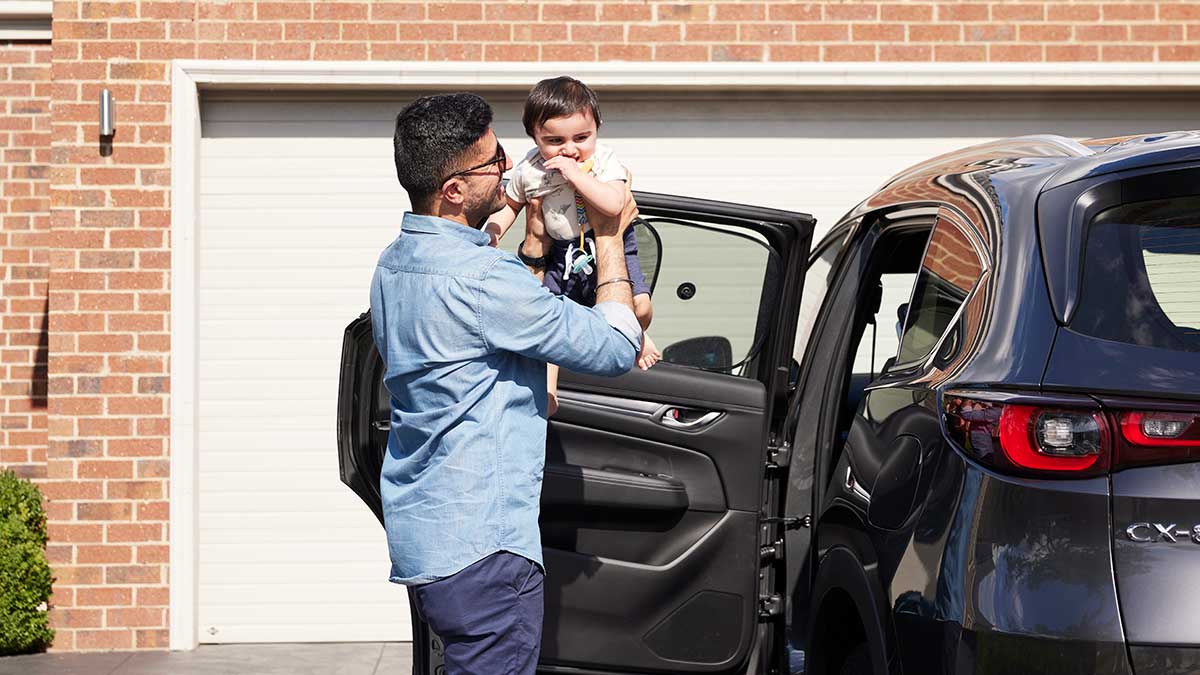 Father in blue shirt lifting small child out of child restraint from Mazda SUV parked in driveway.