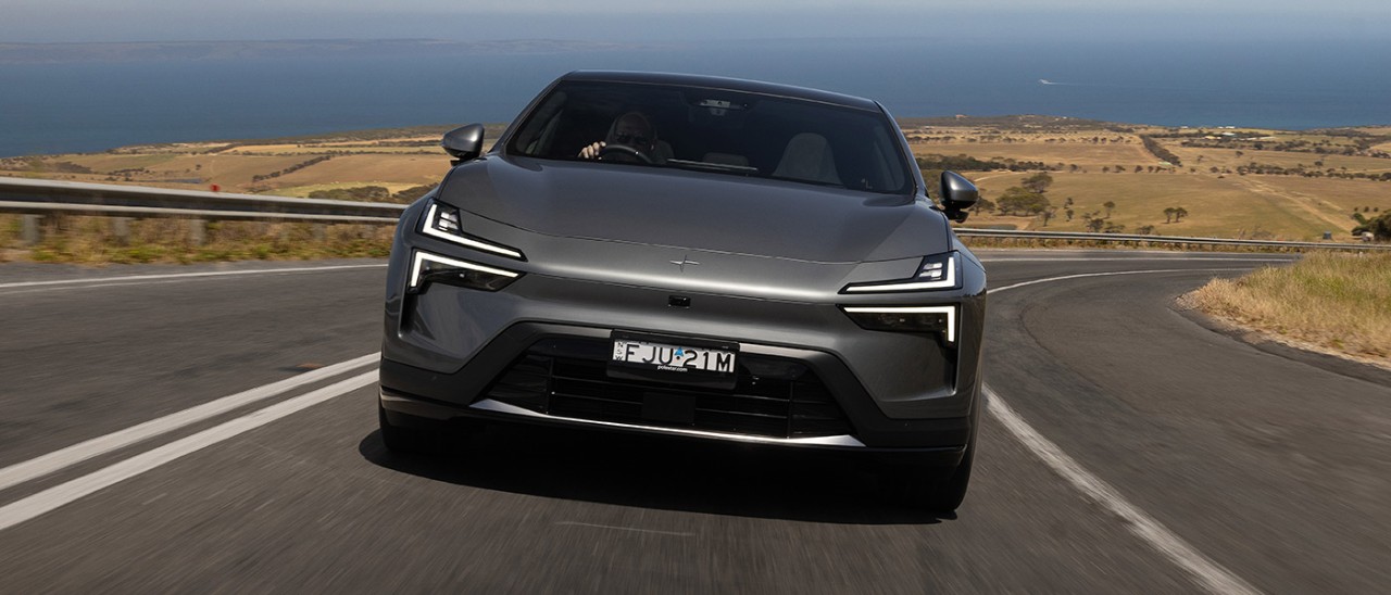 Grey metallic Polestar 4 SUV driving uphill on rural highway with ocean in background